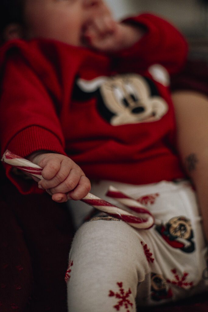 Nahaufnahme einer Hand eines Babys (Mädchen), wie sie eine Zuckerstande festhält, im Hintergrund ein Mickey Maus Pullover, bei einem Babyshooting in Kaiserslautern, Pfalz, an Weihnachten